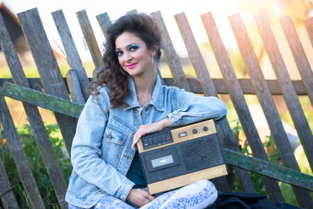 A beautiful country girl in bright clothes sits on a wooden bench with a cassette recorder. Woman in the style of the 90s.の写真素材