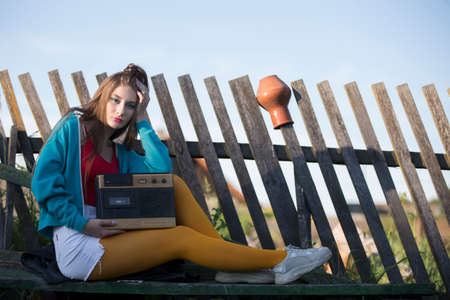 A beautiful country girl in bright clothes sits on a wooden bench with a cassette recorder. Woman in the style of the 90s.の写真素材