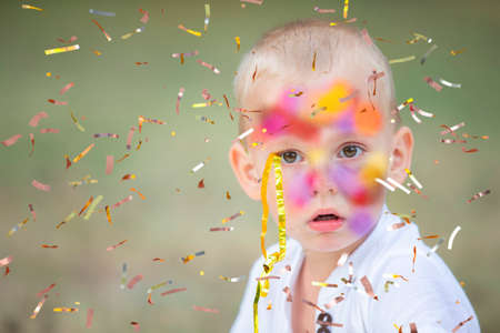 A funny little boy with a face smeared with paints looks at the flying confetti. The child is at the party.の写真素材