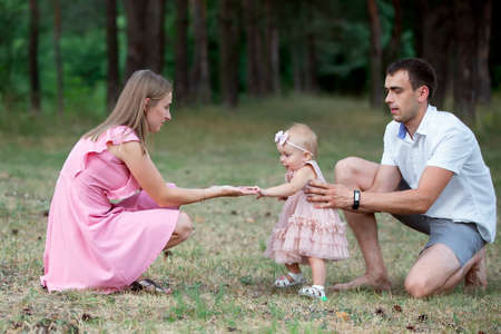 Husband, wife and their little daughter for a walk in the park. Happy dad and mom teach a one-year-old baby to walk.の写真素材