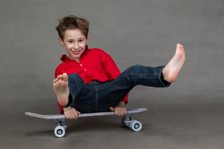 Funny little boy sitting on a vintage skateboard barefoot on a gray background.