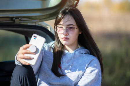 A beautiful girl sits in a car and looks at the phone screen. Taking a selfie in nature or...