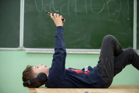 The child is resting at school. A schoolboy boy lies in headphones on a school desk and looks into a...