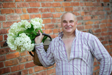 Elderly male florist. A man holds a watering can with a bouquet of flowers against a brick wall. Passion for the garden.の写真素材