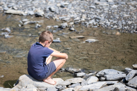 Little boy sitting on rocks with his back looking at the mountain lake.の写真素材