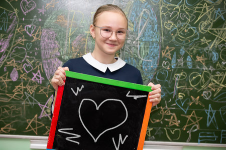 A schoolgirl of middle school age in glasses holds a slate board on which a heart is drawn.