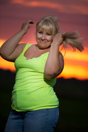 A plump middle-aged woman poses in jeans and a bright t-shirt against the background of the evening...