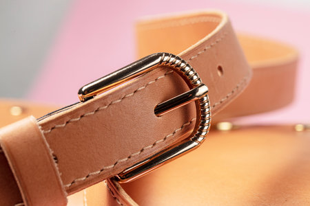 Part of an expensive brown leather item with stitching and a metal buckle on a pink background.