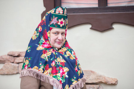 An elderly Russian woman in a traditional headdress, kokoshnik, poses against the backdrop of a village house.の写真素材