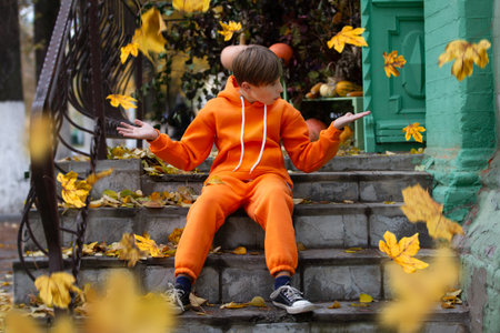 A boy in orange clothes sits on the steps and looks at the yellow flying autumn leaves. Fall has come.の写真素材