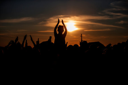 An unrecognizable group of people waves their hands at a concert or public event against the backdrop of the setting sun. Night music festival.の写真素材