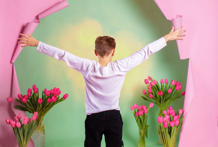 The boy stands with his back to a pink paper background with spring tulips.