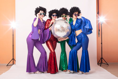 Three female pop stars in neon clothes pose in front of a silver ball in a studio. Group of girls in...