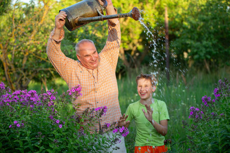 A grandfather waters flowers with a watering can while his grandson watches and laughs. The scene is peaceful and serene, grandfather and grandson enjoying the simple pleasure of tending to their garden.の写真素材
