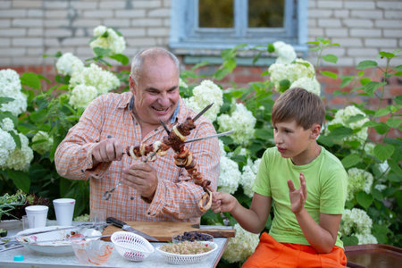 A grandfather and his grandson are happily eating shashlik from a skewer and chatting in the yard of a village house. A man and a boy are eating together.の写真素材