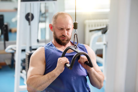 A man works out in the gym, lifting weights with a barbell. He is wearing a blue tank top and is...