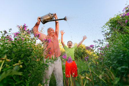 A man and a boy are standing in a garden, the man is holding a watering can and the boy is laughing. The scene is carefree and joyful as the two men enjoy time together in the garden.の写真素材