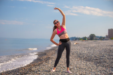 An adult woman in a sports bra and leggings does bending exercises in the coastal area.の写真素材