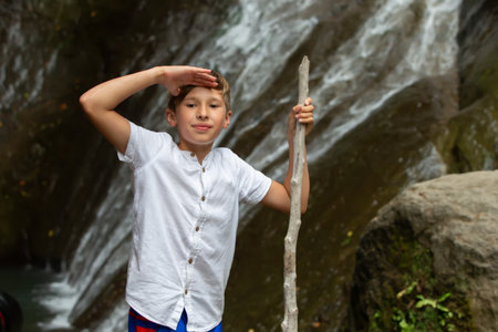 A tourist boy in a white shirt with a stick against the background of a blurred waterfall with a mountain.の写真素材