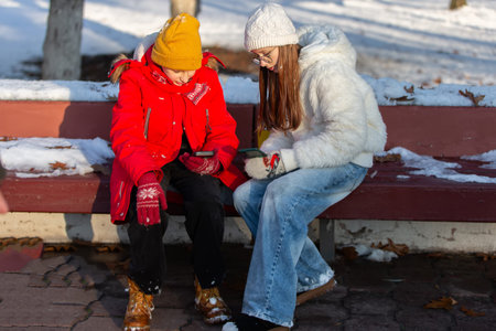 A boy and a girl in winter clothes are sitting on a bench with smartphones.