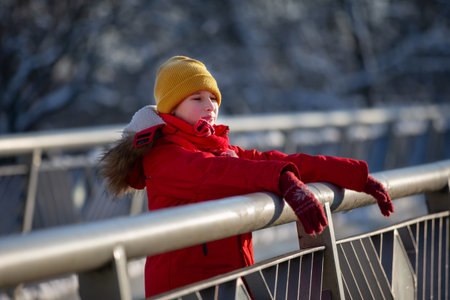 A boy in a red jacket and a yellow hat stands on a bridge and does not look at the camera.