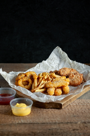 Fast Food. Salty Snacks. French Fries, Potato Chips, Onion Rings, Cheese Sticks, Nuggets wiht Sauce on Rustic Wooden Table on Dark Background. Concept Food For Partiesの写真素材