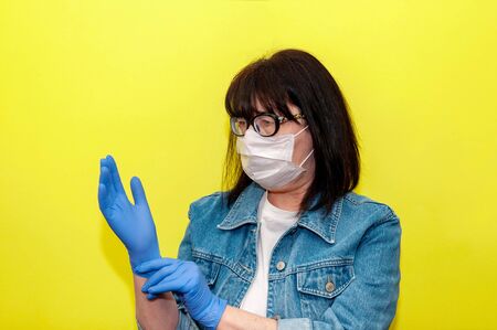 COVID-19. Senior Woman in Protective Face Mask Puts On Medical Gloves on Yellow Background. Anti-Coronavirus Concept.の写真素材