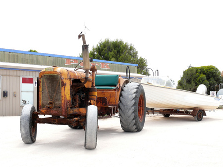 Yalata, South Australia - December 4, 2007: Very old tractor with a trailer. Motor boat on the trailer. Architectural structures.のeditorial素材