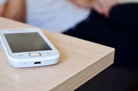 white mobile phone lies on the edge of the table against the background of a seated man horizontal orientationの写真素材