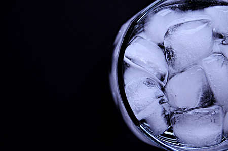 Half a glass of full drinking water with ice cubes close up on a black background view from above...