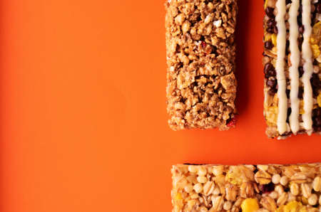 Three granola bars lie on an orange background on the right side of the photo view from above with a...