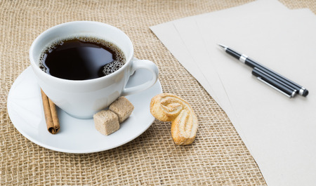 Cup of black coffee with foam, cookie, brown sugar, craft paper, pen and cinnamon stick on sackcloth backgroundの写真素材