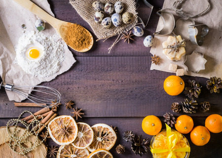 Christmas background frame with tangerines, dried oranges, cinnamon sticks, anise stars, cinnamon on spoon, cookie molds, flour with egg on paper. Rustic brown wooden background. Top viewの写真素材