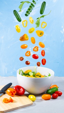 Levitating mixed vegetables on blue background. Magical cucumber, tomato and paprika with white bowlの写真素材