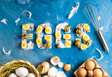 Chicken and quail eggs with word eggs on blue background with basket, feathers, whisk and boxの写真素材