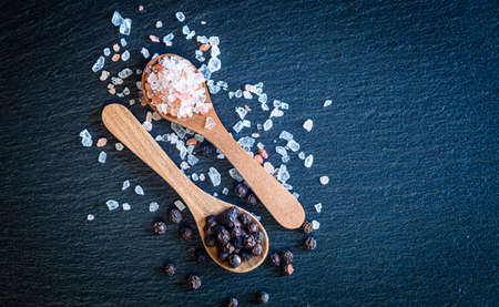 Himalaya pink sea salt and whole black pepper in wooden spoon on dark stone background, close view