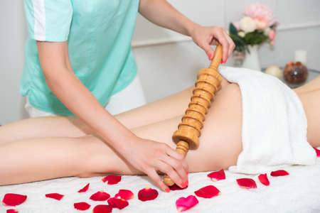 woman massage therapist doing anti-cellulite massage, to a young girl, a wooden roller massager. legs covered with a white towel. on the couch are rose petals. health concept, slim body.の写真素材