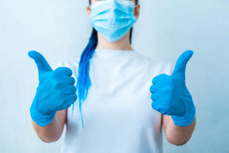 Cropped photo close-up of a girl with blue hair in gloves and a mask showing thumb up, isolated on a light background. class symbolの写真素材