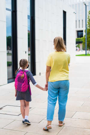 mother leads her daughter to study by the hand. walk together in the park, rear view. girl in uniform, with a school bagの写真素材