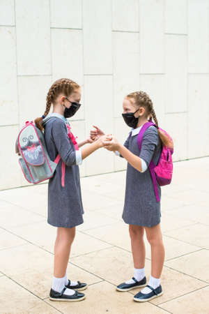 Two elementary school students, in medical masks on the face, walk hand in hand. sisters with school...