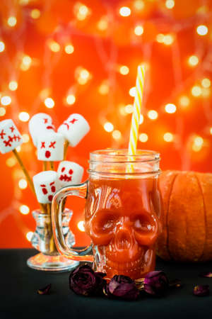 glass mug skull with red tomato juice. a welcome cocktail at a halloween party. marshmello scary faces, ghosts. pumpkin and garland lights, bokeh. orange background.の写真素材