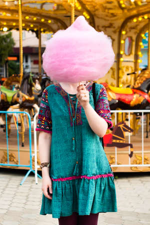 Young beautiful hipster woman eating cotton candy in an amusement park. girl covered her face with...