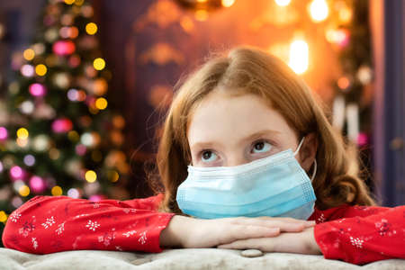 red-haired girl in a medical mask is sad on Christmas eve. garland, bokeh, holiday. flu prevention, coronavirus, covid-19, isolation, stay at homeの写真素材