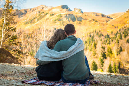 Autumn Walk, mountains. Rear view of couple sits and admires the fall forest. Man and woman in warm clothing and scarfの写真素材
