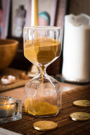 Photo for antique hourglass counts down the time. coins, money and candles on the table. sand falls in hours, dries up. - Royalty Free Image