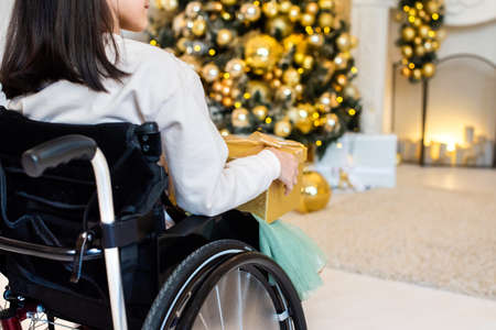 little girl in a wheelchair near the Christmas tree. disabled person holding yellow, golden New Years giftの写真素材