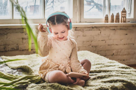 a little girl sits on the bed in headphones. baby recklessly plays games and listens to musicの写真素材