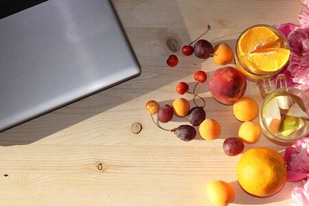 Apricot and orange juice, apricots, grapes on a sunny table next to a laptop, copy space, top view, flat lay. Summer drinks and fruits during a break at work with colorful peony branches, selective focusの写真素材