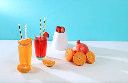 Modern still life with citrus, orange and strawberry juice and oranges, pomegranate and strawberries on stand and podiums on blue background with long hard shadows, minimal detox diet concept, summer vitamin drinks, selective focusの写真素材