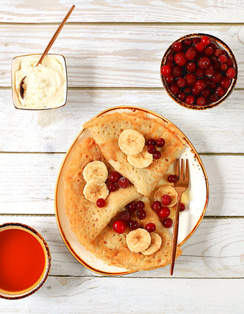Delicious Russian pancakes with soft curd cheese, bananas, cranberries and honey on a wooden table....